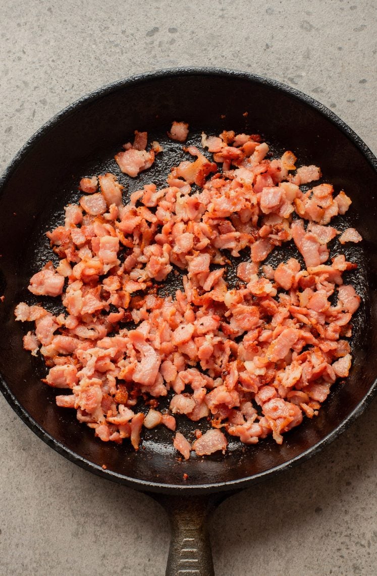 Chopped bacon pieces cooking in a black cast iron skillet on a speckled gray countertop.