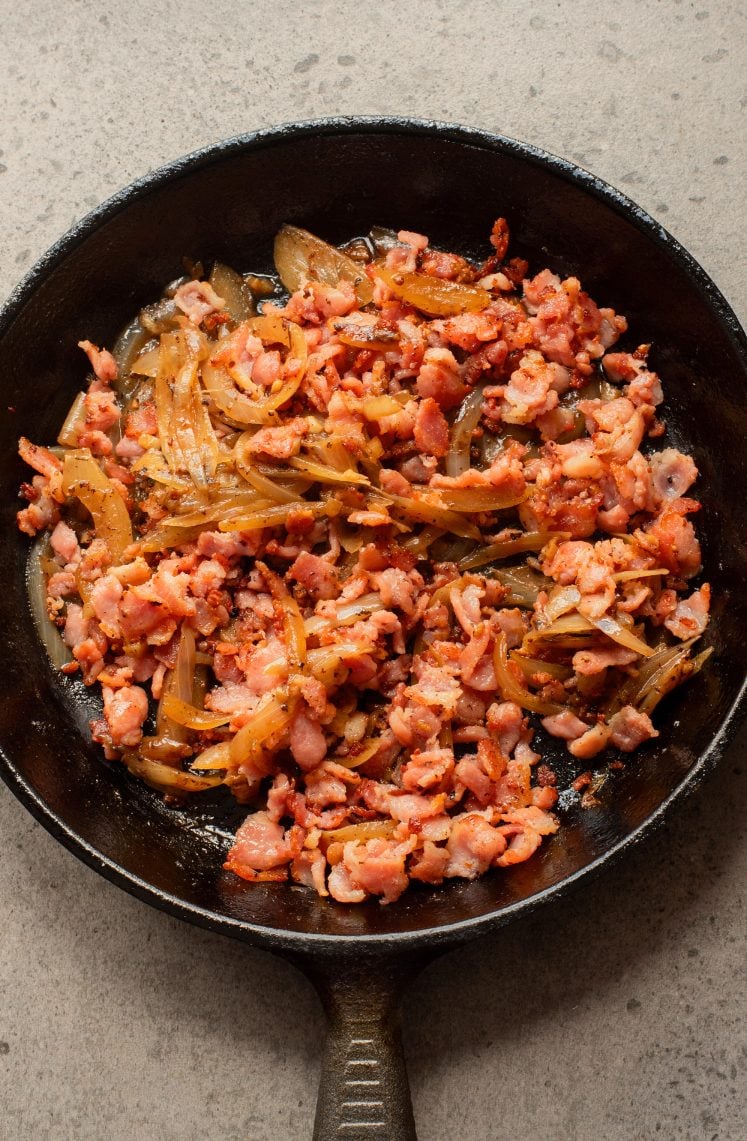 A cast iron skillet containing sautéed chopped bacon and sliced onions on a light gray surface.