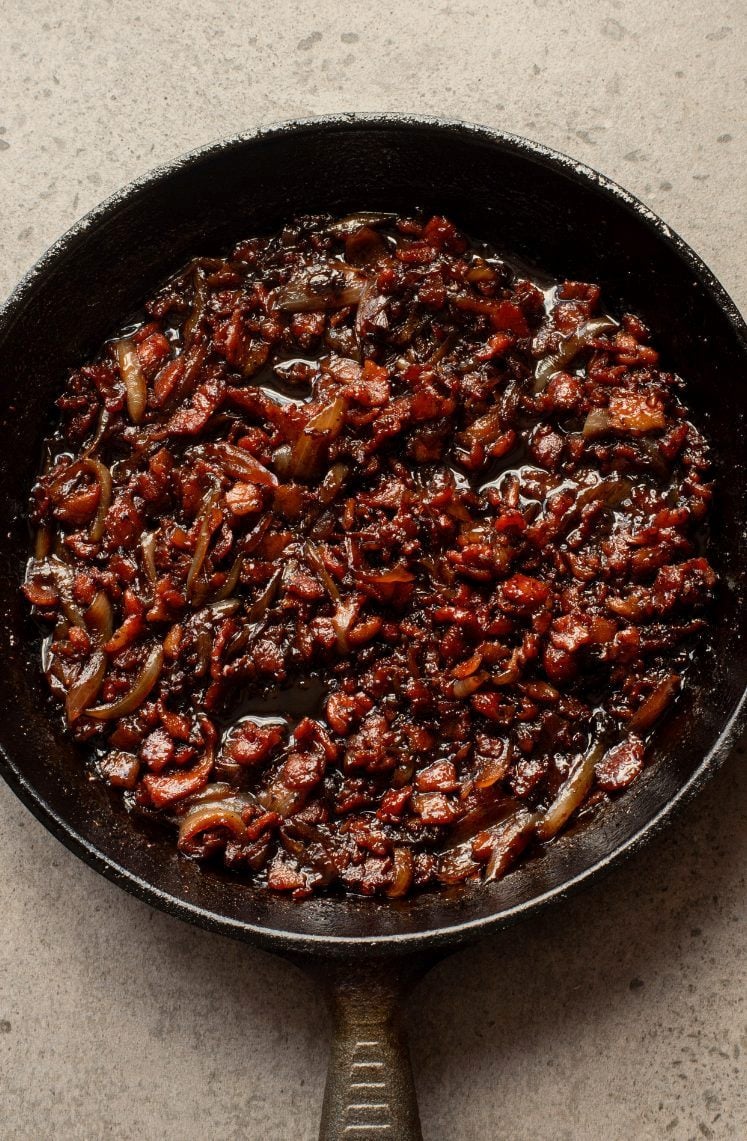 A cast iron skillet filled with cooked bacon and onions in rendered fat, photographed from above on a light-colored surface.