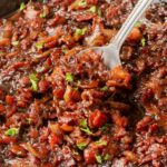Rich, caramelized bacon onion jam being scooped from a skillet, topped with fresh parsley.