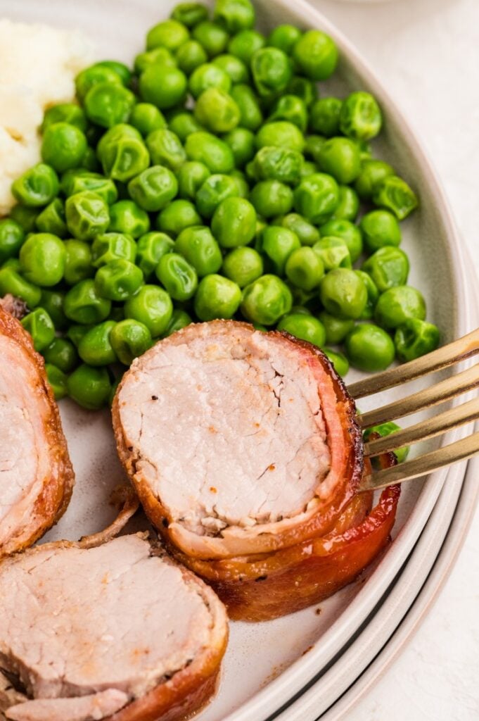 A plate with bacon-wrapped pork tenderloin slices, green peas, and mashed potatoes, with a fork holding a piece of the pork.