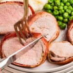 A fork and knife cut into bacon-wrapped pork medallions on a plate with mashed potatoes and green peas.