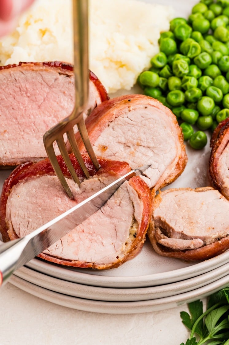 A fork and knife cut into bacon-wrapped pork medallions on a plate with mashed potatoes and green peas.