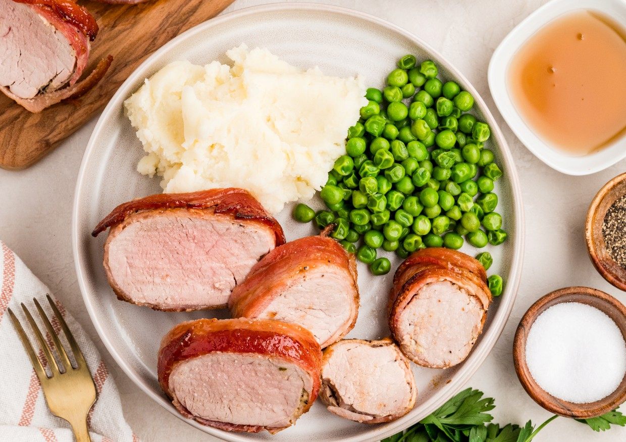 A plate with bacon-wrapped pork tenderloin slices, mashed potatoes, and green peas. Small bowls of sauce, pepper, and salt are nearby.