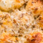 Close-up of a baked breakfast casserole topped with melted cheese, browned sausage, and golden biscuit pieces in a white baking dish.