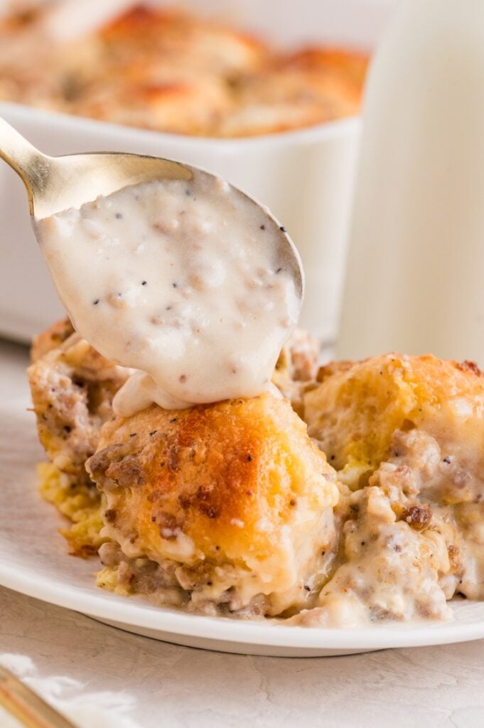 A spoon pours creamy sausage gravy over a serving of biscuits on a white plate, with a casserole dish and bottle of milk in the background.