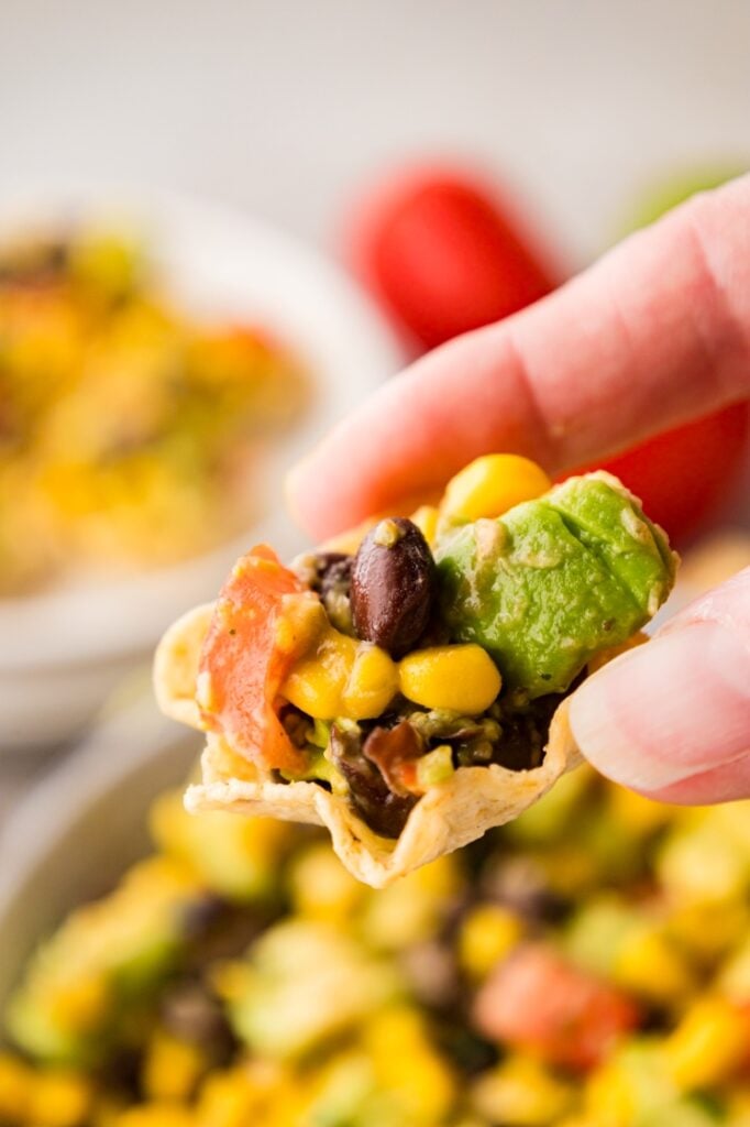 A hand holds a tortilla chip topped with avocado, black beans, corn, and diced tomatoes, with a blurred bowl of similar dip in the background.