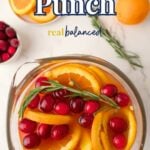 Overhead view of a glass pitcher filled with Christmas punch, garnished with orange slices, cranberries, and rosemary. Whole oranges, cranberries, and greenery are nearby.