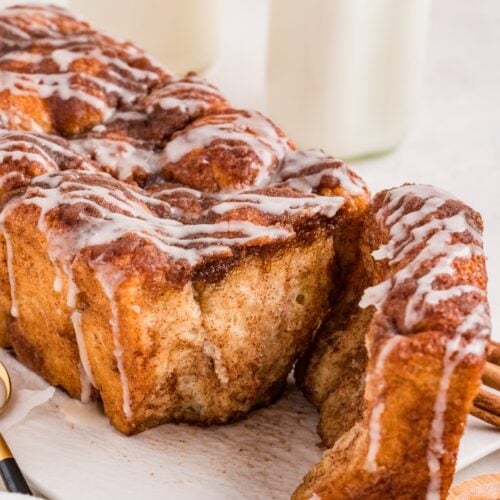 A loaf of cinnamon pull-apart bread with icing drizzled on top, with one piece partially pulled away; two bottles of milk are in the background.