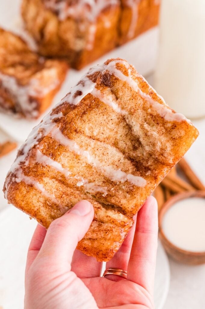 A hand holds a square piece of cinnamon pull-apart bread topped with white icing, with more bread and a glass of milk in the background.