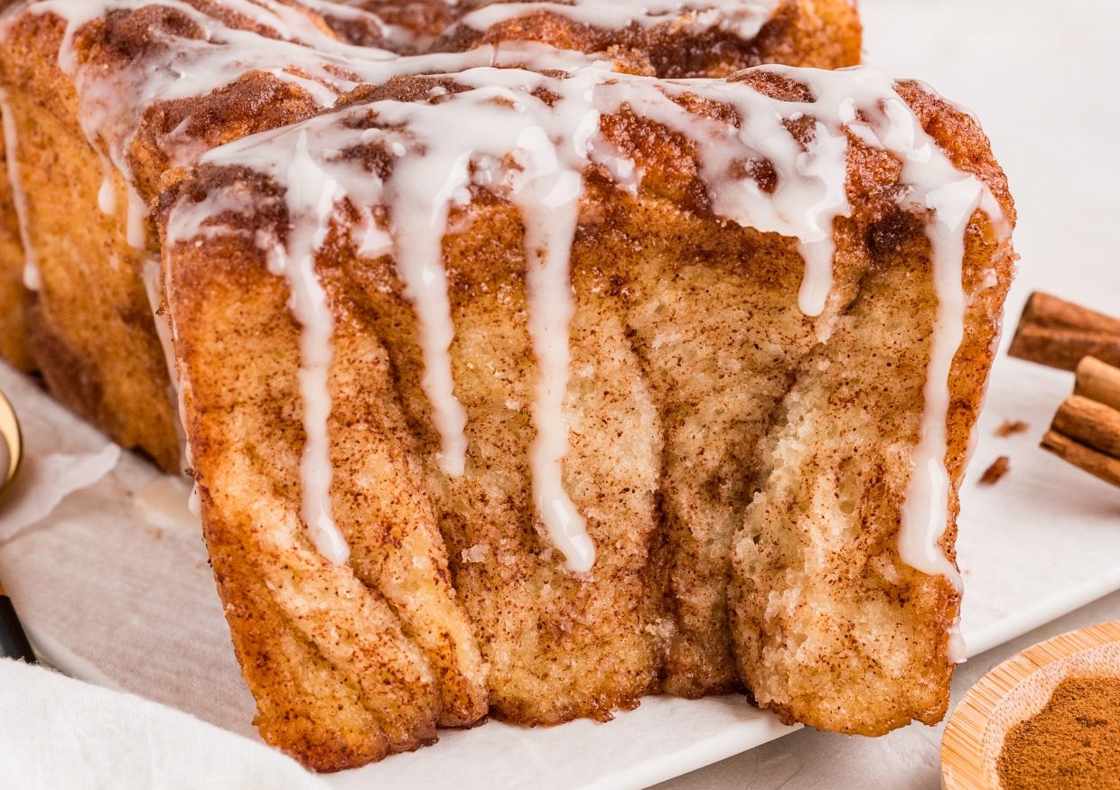 A loaf of pull-apart cinnamon bread drizzled with white icing, with visible swirls of cinnamon and a soft texture, sits on a piece of parchment paper.