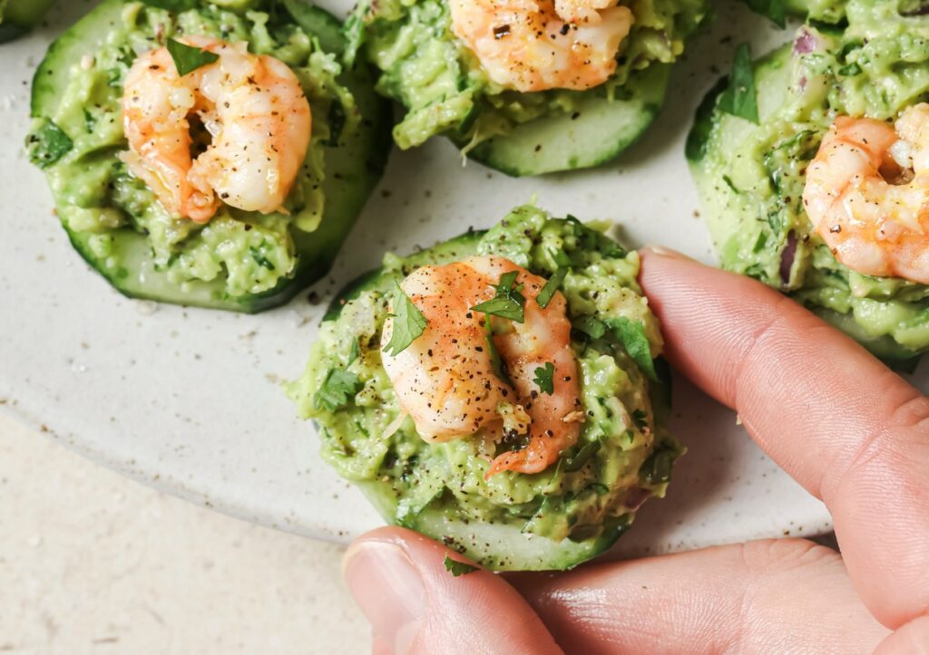 A hand picking up a cucumber slice topped with mashed avocado and a cooked shrimp, garnished with herbs and black pepper, from a plate.