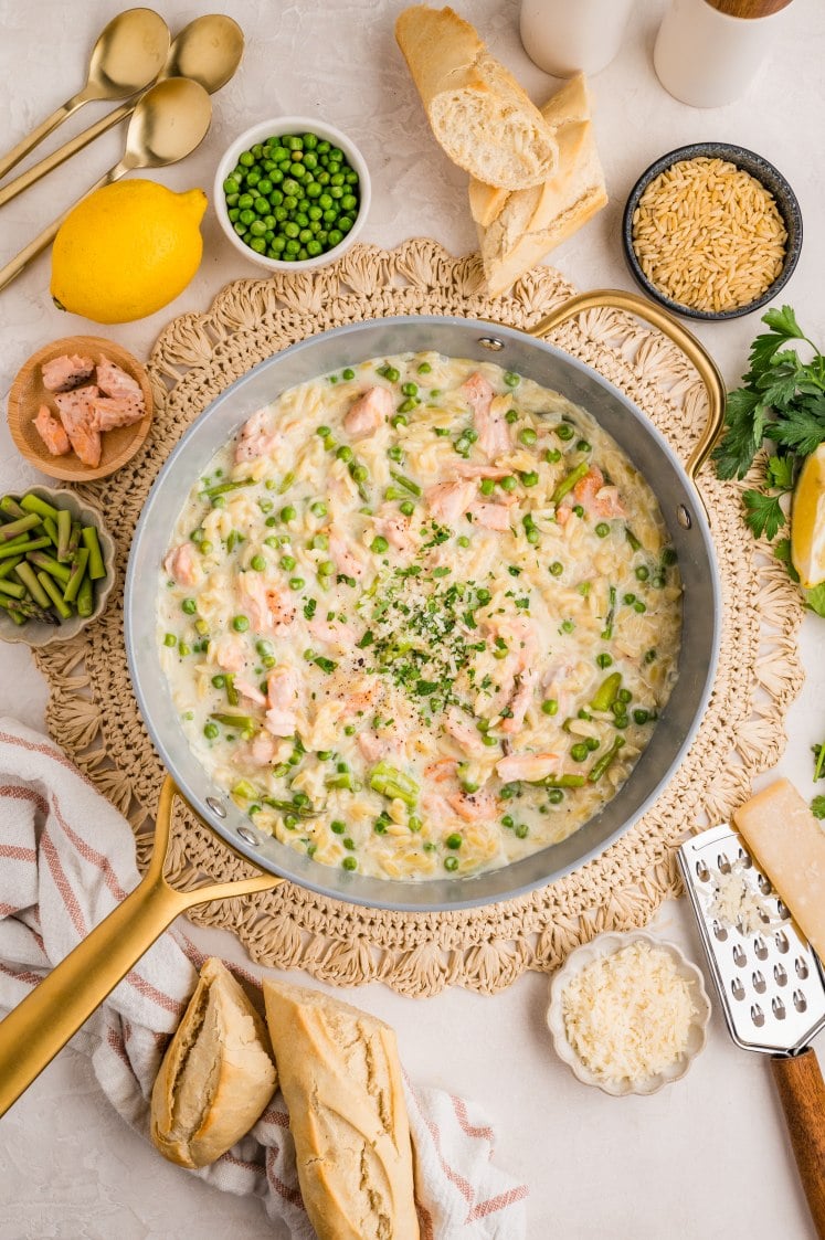 Creamy salmon orzo finished in a skillet with peas and asparagus, garnished with herbs.