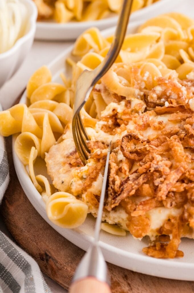 A plate of egg noodles served with a creamy chicken dish topped with crispy fried onions, being cut with a knife and fork.