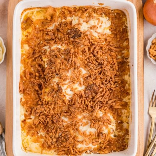 A baked casserole topped with crispy fried onions in a white rectangular dish on a wooden board, surrounded by onions, utensils, and a striped cloth.