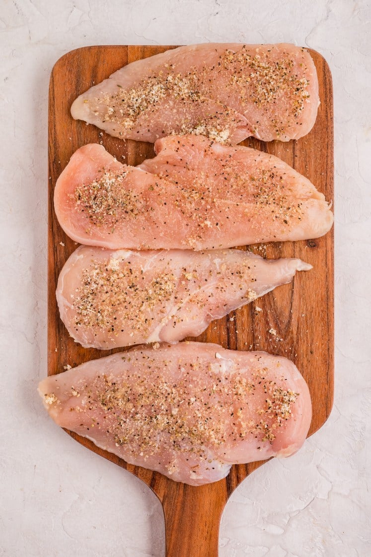 Raw chicken breasts seasoned with herbs and spices on a wooden cutting board.