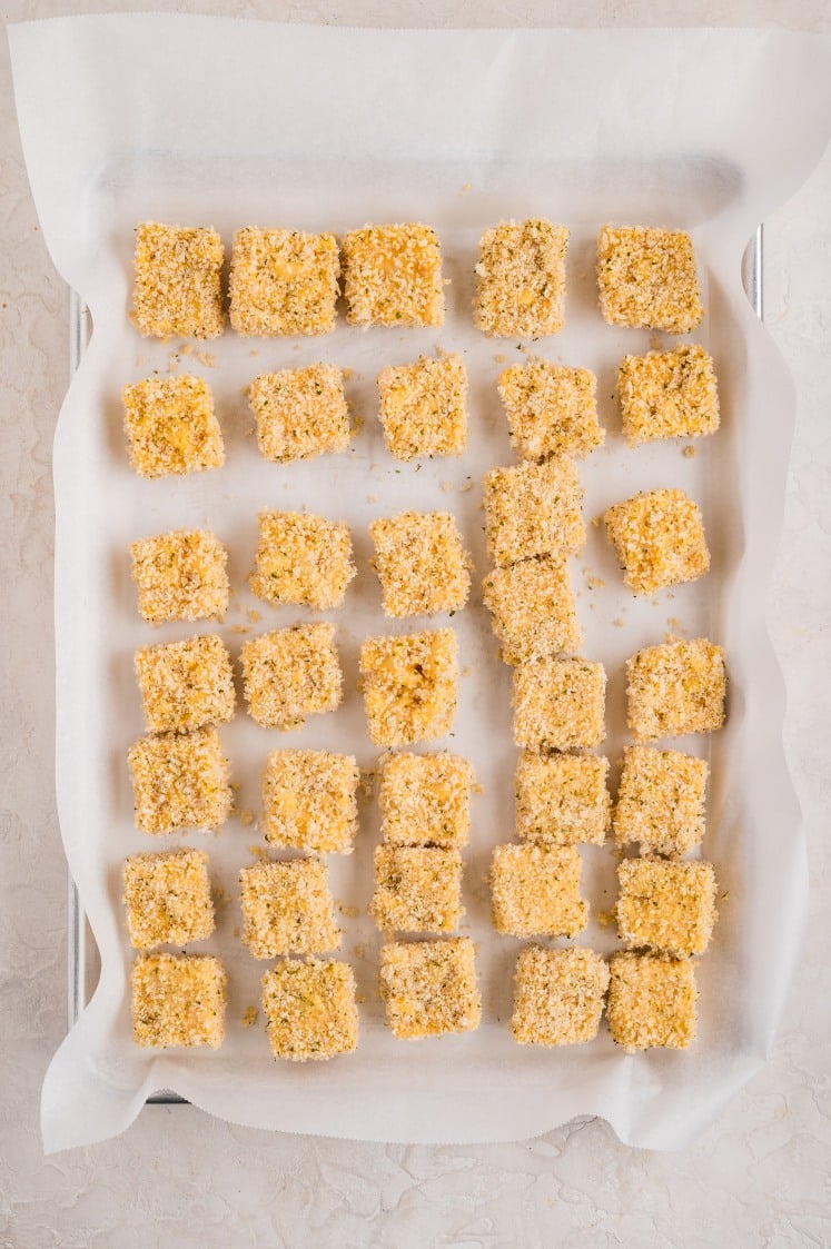 Breaded mac and cheese cubes arranged on a parchment-lined baking sheet.