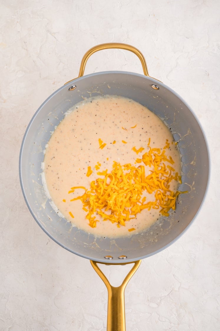 Shredded cheddar cheese added to creamy sauce in a saucepan.