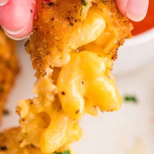 Close-up of a hand pulling apart a breaded, fried macaroni and cheese bite, showing melted cheese inside.