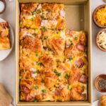 A baking pan filled with a croissant breakfast casserole topped with ham, cheese, and chopped green onions, surrounded by ingredients like eggs, cheese, and green onions.