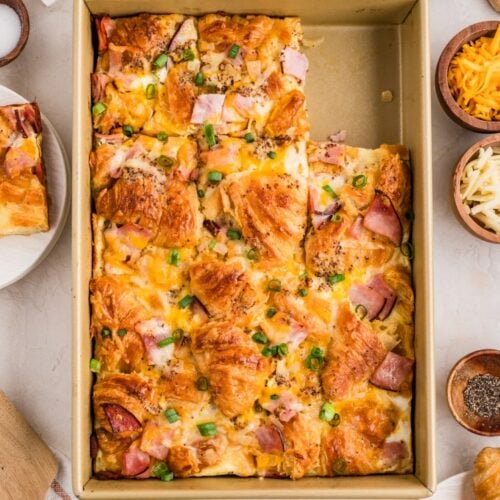 A baking pan filled with a croissant breakfast casserole topped with ham, cheese, and chopped green onions, surrounded by ingredients like eggs, cheese, and green onions.