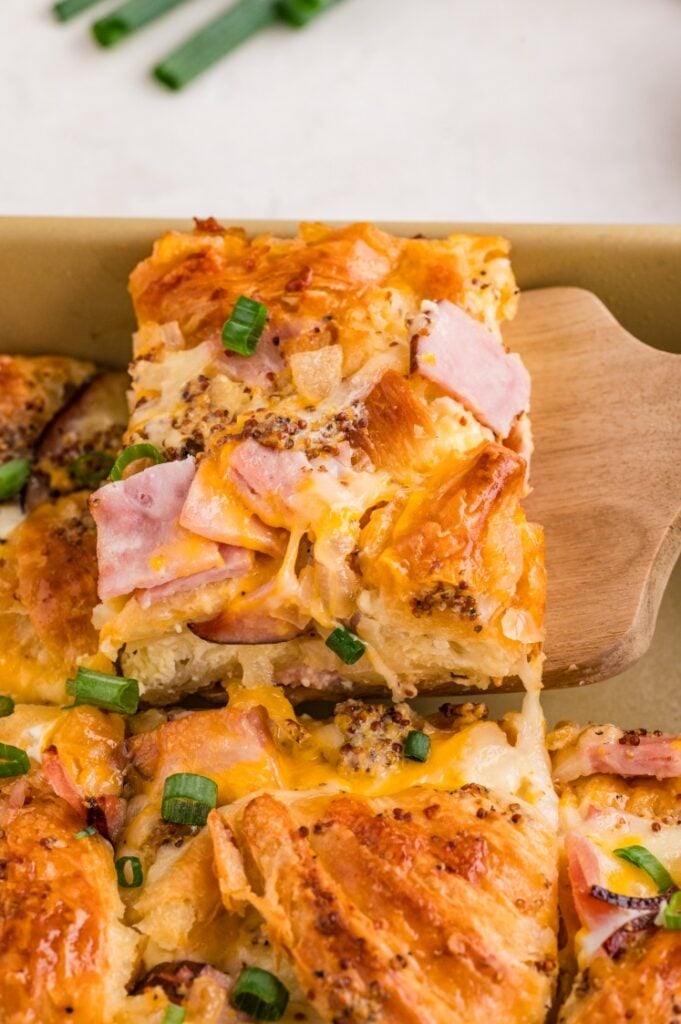 A slice of baked casserole with croissants, ham, melted cheese, and chopped green onions being lifted from a pan with a spatula.