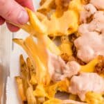 A hand lifts cheesy loaded fries topped with melted cheese and a creamy sauce from a tray lined with parchment paper.