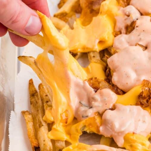 A hand lifts cheesy loaded fries topped with melted cheese and a creamy sauce from a tray lined with parchment paper.