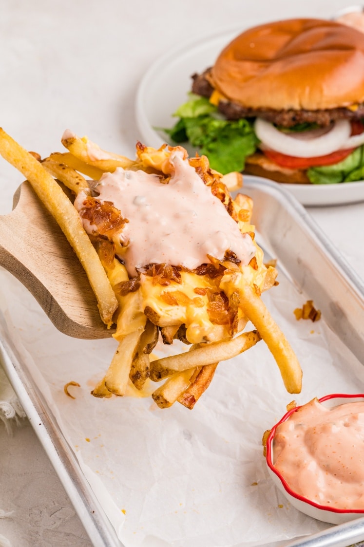 French fries covered in melted cheese, grilled onions, and sauce are served on a tray, with a cheeseburger and dipping sauce in the background.