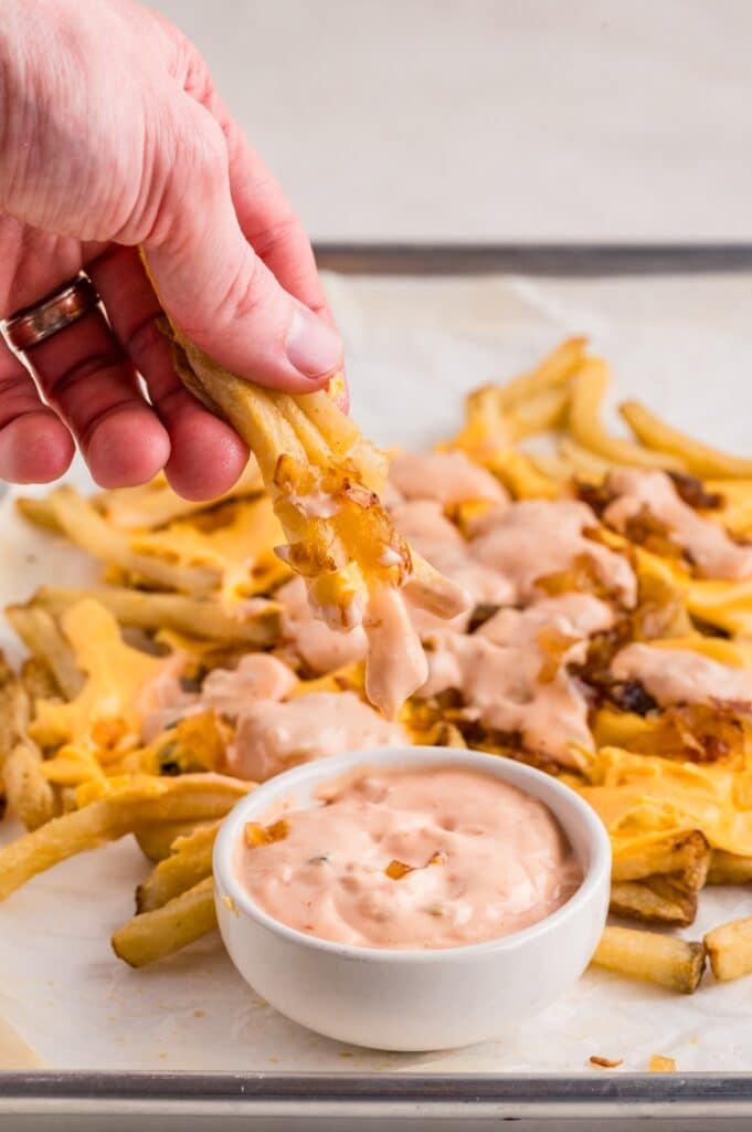 A hand dips cheese-covered fries topped with grilled onions into a bowl of pink sauce, with more loaded fries on parchment paper in the background.