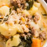 A close-up of a bowl of soup containing ground meat, potatoes, kale, white beans, carrots, and topped with grated cheese.