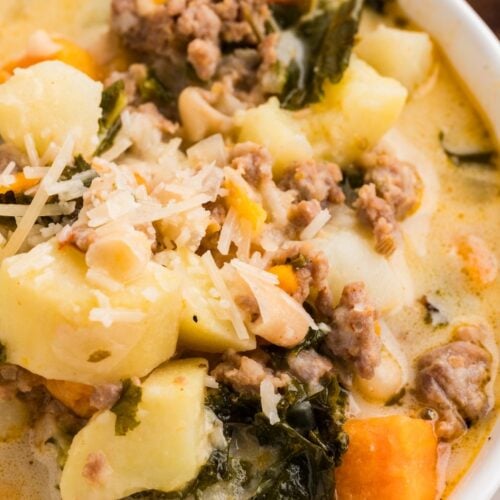 A close-up of a bowl of soup containing ground meat, potatoes, kale, white beans, carrots, and topped with grated cheese.