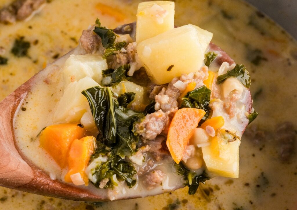 A wooden spoon holding a serving of creamy soup with potatoes, carrots, kale, ground meat, and white beans.