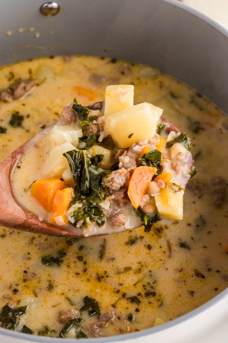 A wooden spoon holds a serving of creamy soup with sausage, potatoes, sweet potatoes, kale, and herbs above a pot of the same soup.