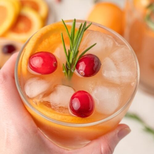 A hand holds a glass filled with ice, orange-colored beverage, orange slice, cranberries, and a sprig of rosemary.