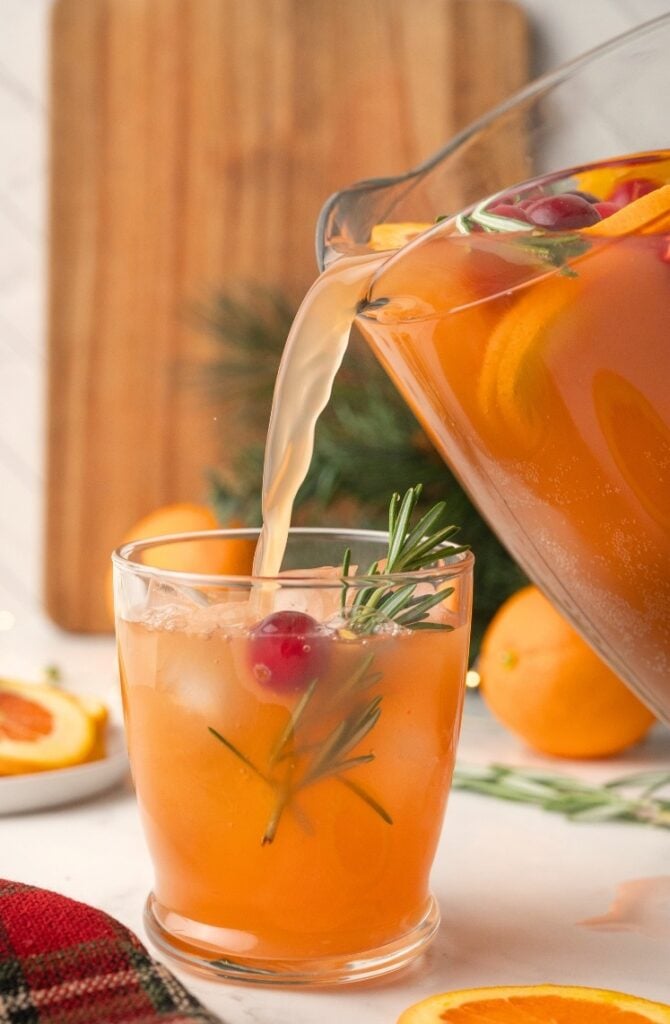 A glass is filled with an orange-colored drink from a pitcher containing orange slices, cranberries, and rosemary; ice and garnishes are in the glass.
