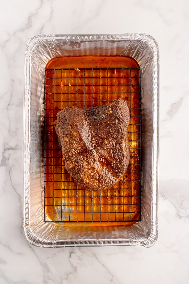 A seasoned piece of meat sits on a wire rack inside a foil tray with juices collected at the bottom, on a marble surface.