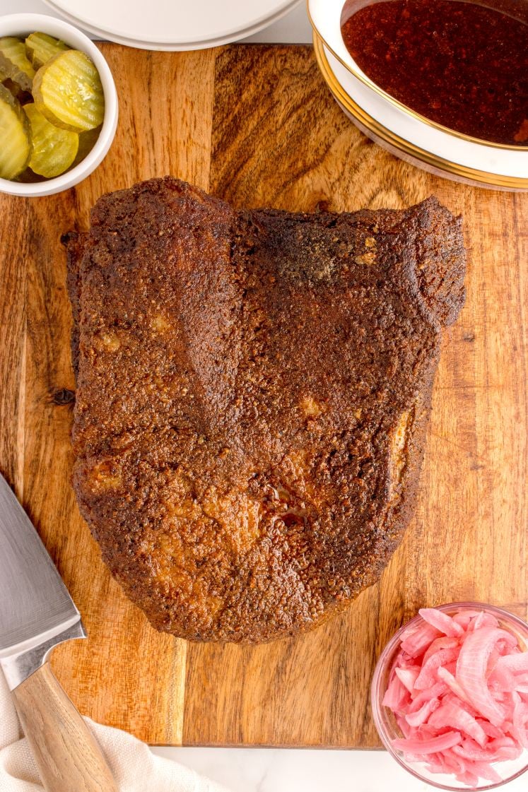 A smoked brisket sits on a wooden cutting board, surrounded by pickles, sliced onions, barbecue sauce, and a large knife.