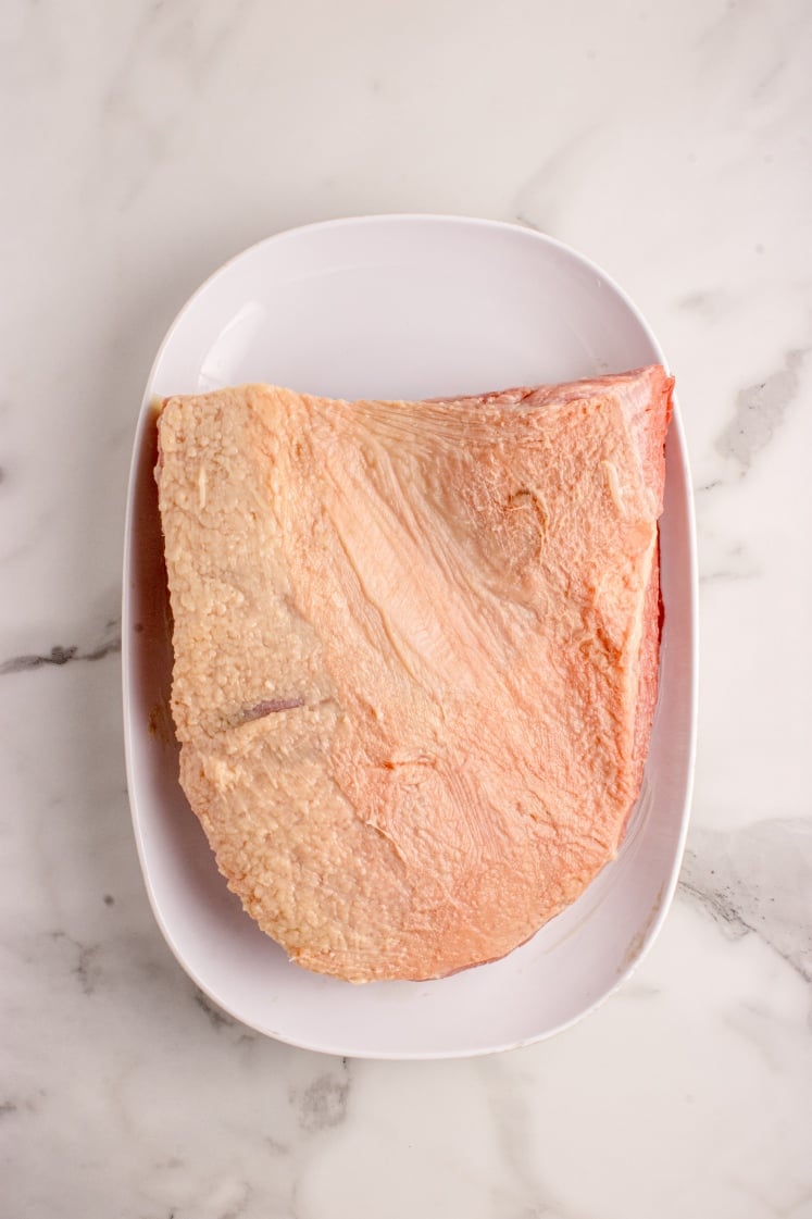 Raw brisket with a thick fat cap resting on a white oval plate against a marble countertop.