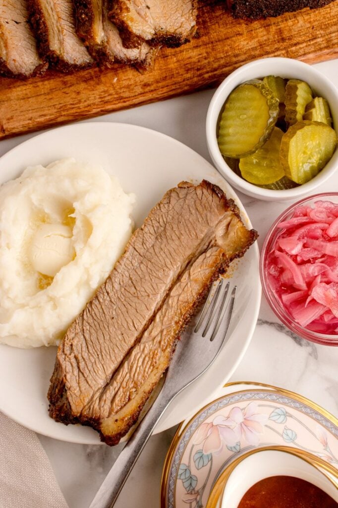 A plate with a slice of brisket, mashed potatoes with butter, and a fork. Nearby are bowls of pickles and pickled onions, with sliced brisket on a wooden board in the background.