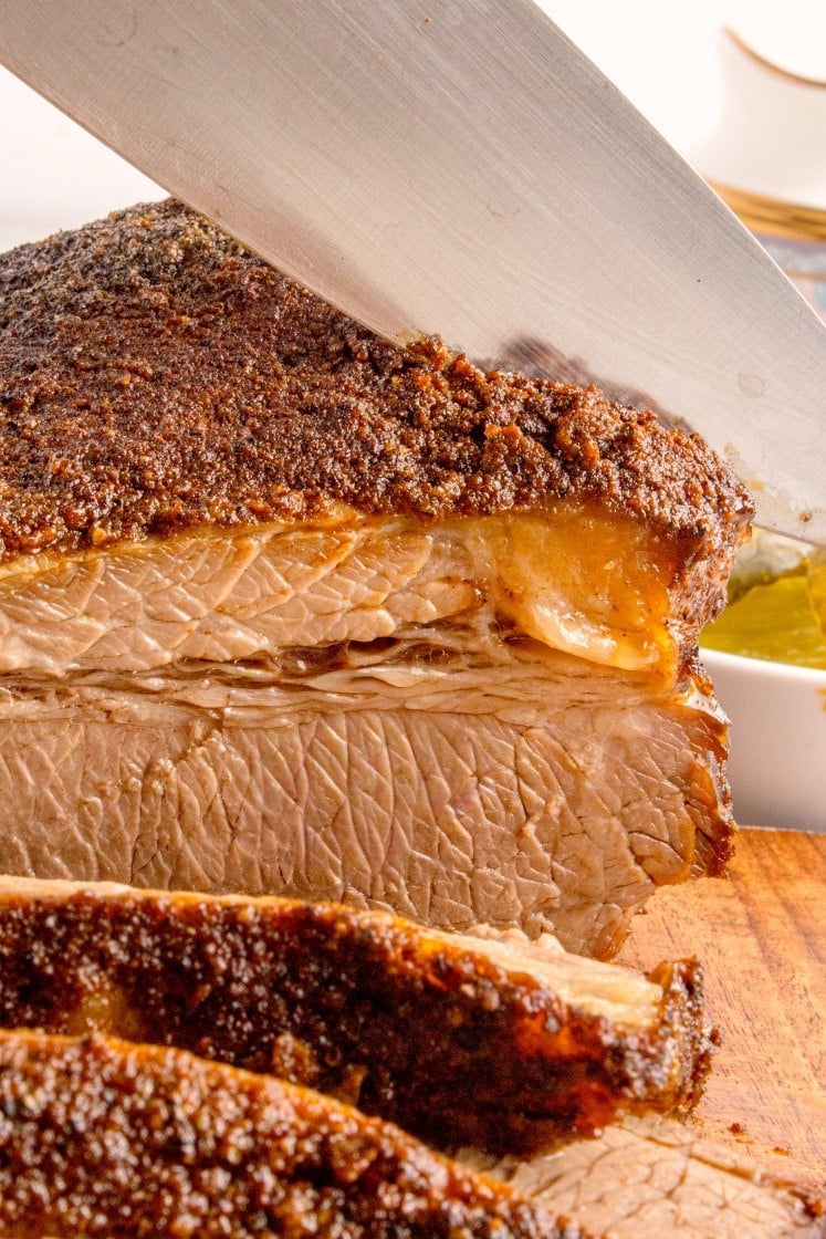 A close-up of a knife slicing through a seasoned, cooked brisket on a wooden cutting board.