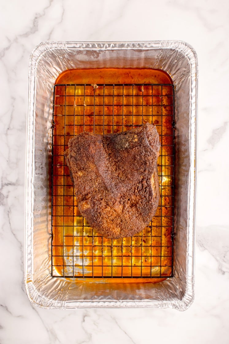 Cooked oven-baked brisket resting on a wire rack inside a foil pan, surrounded by rendered juices.