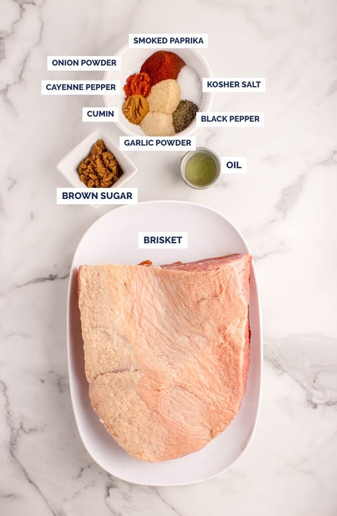Uncooked brisket on a white plate with labeled bowls of brown sugar, onion powder, cayenne pepper, cumin, smoked paprika, kosher salt, black pepper, garlic powder, and oil.