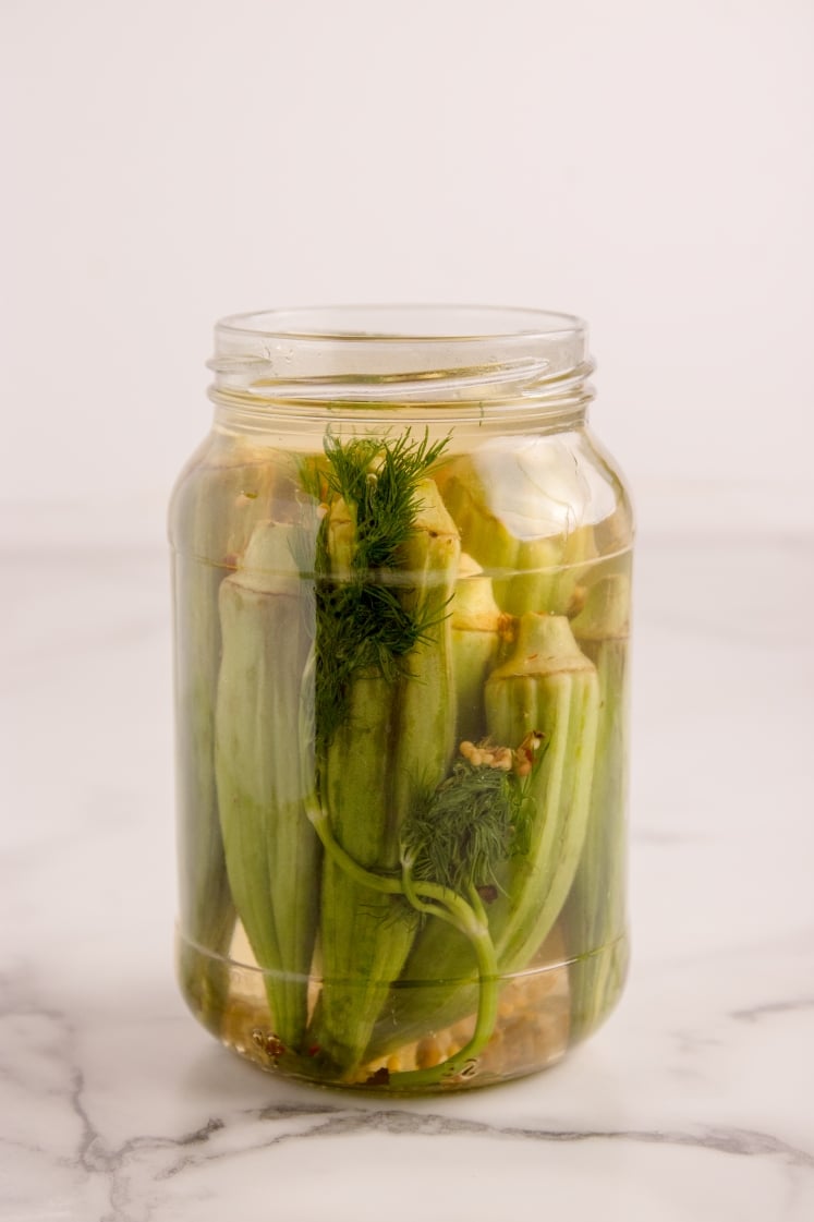 Glass jar of pickled okra in brine with garlic, dill, and spices.