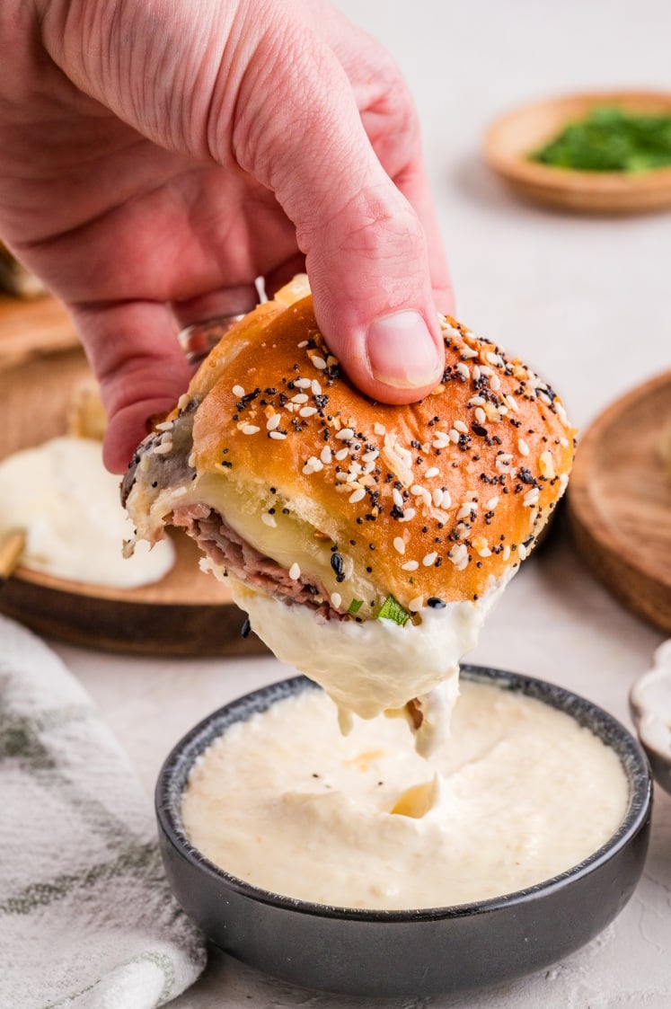 A hand dips a sandwich with an everything bagel bun, roast beef, and cheese into a bowl of white sauce.