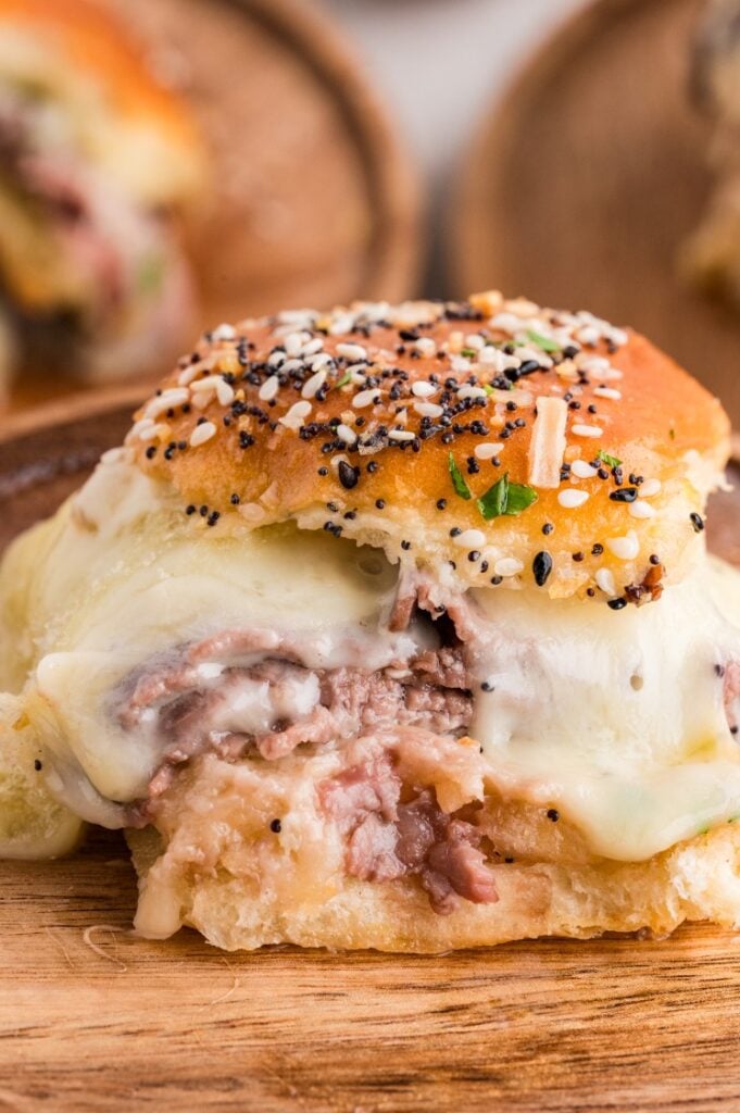 Close-up of a sandwich slider with melted cheese, shredded meat, and an everything-seasoned bun on a wooden surface.