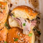 Close-up of baked sliders with golden buns, melted cheese, roast beef, and sprinkled sesame and poppy seeds, garnished with fresh herbs.