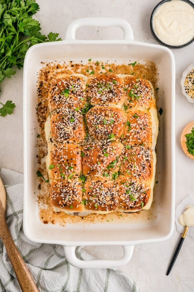 A white baking dish filled with twelve golden slider buns topped with seeds and chopped herbs, surrounded by various small bowls and kitchen utensils on a countertop.