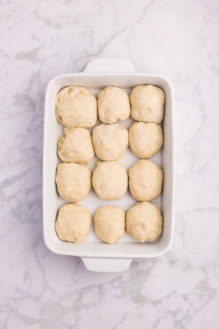 Twelve shaped dough balls arranged evenly in a white baking dish before rising.