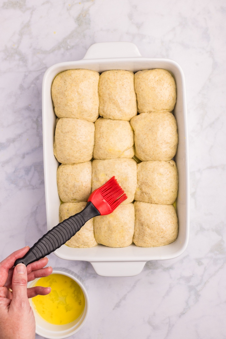 A hand uses a red pastry brush to coat risen dough balls in a baking dish with melted butter.